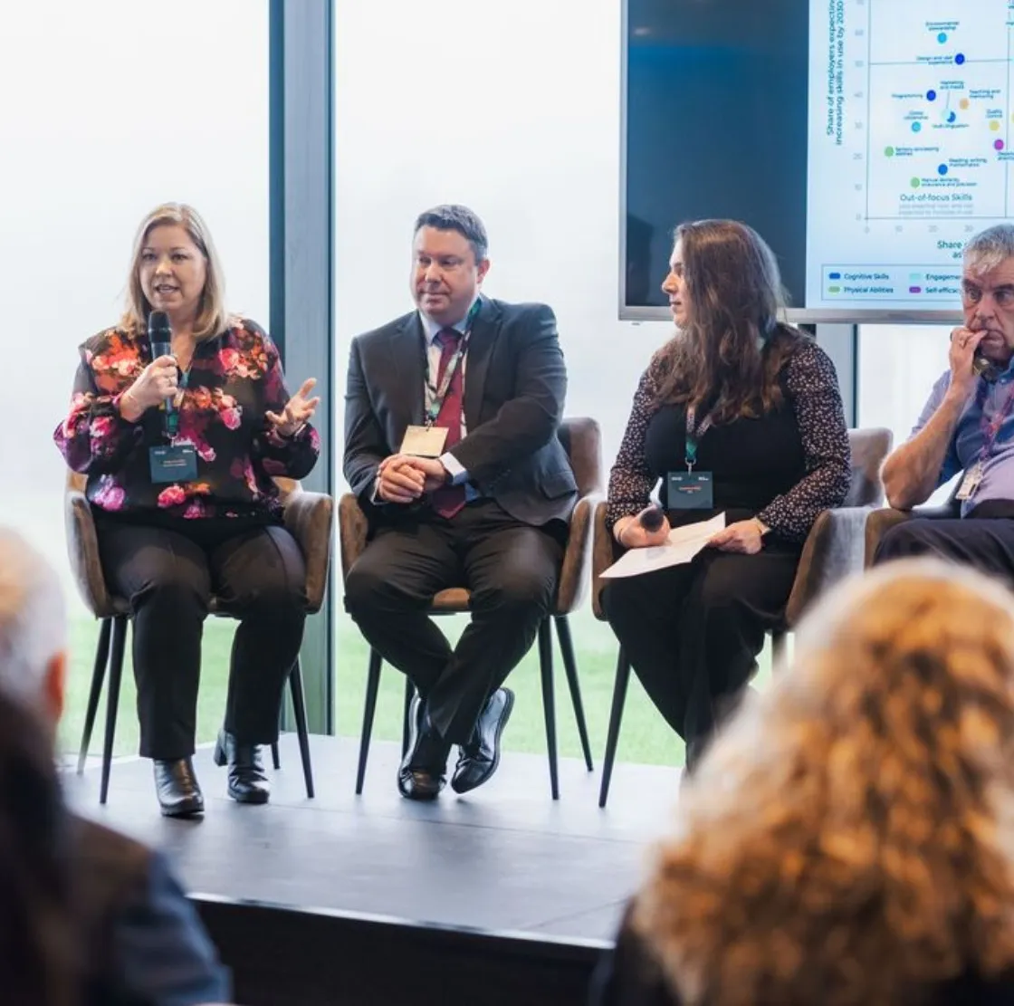 Four panelists seated on chairs engaged in a discussion, with one woman speaking into a microphone and a large screen displaying a chart behind them.
