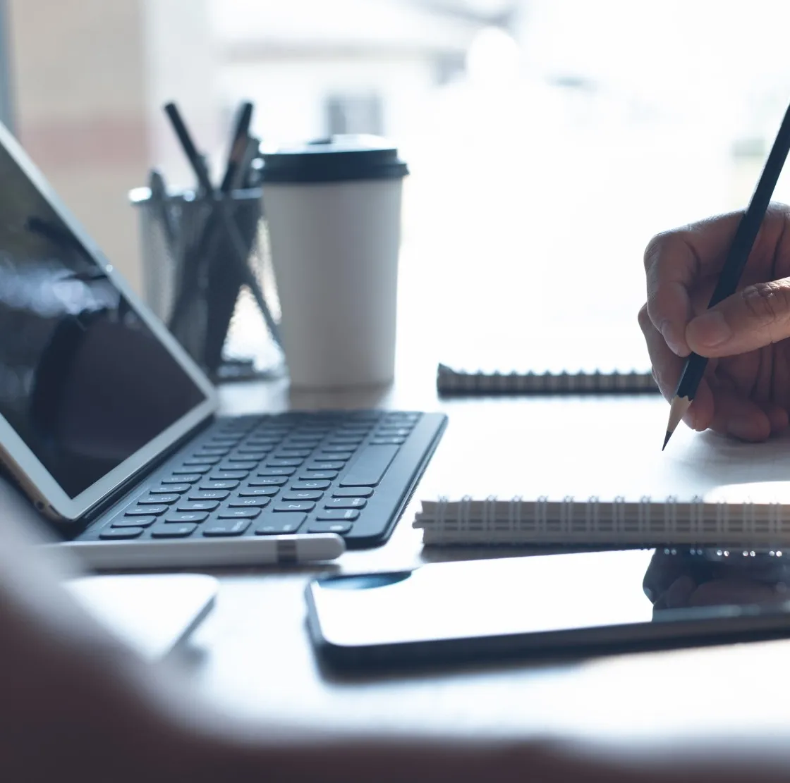 Hand holding a pencil poised over a notebook beside a tablet with keyboard, a smartphone, and a coffee cup on a desk.