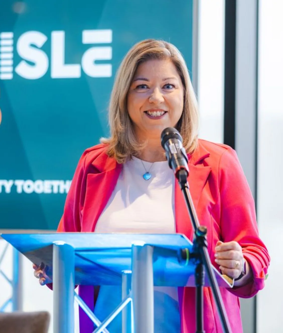 Woman in a pink blazer speaking at a podium with a microphone in front of a green background.