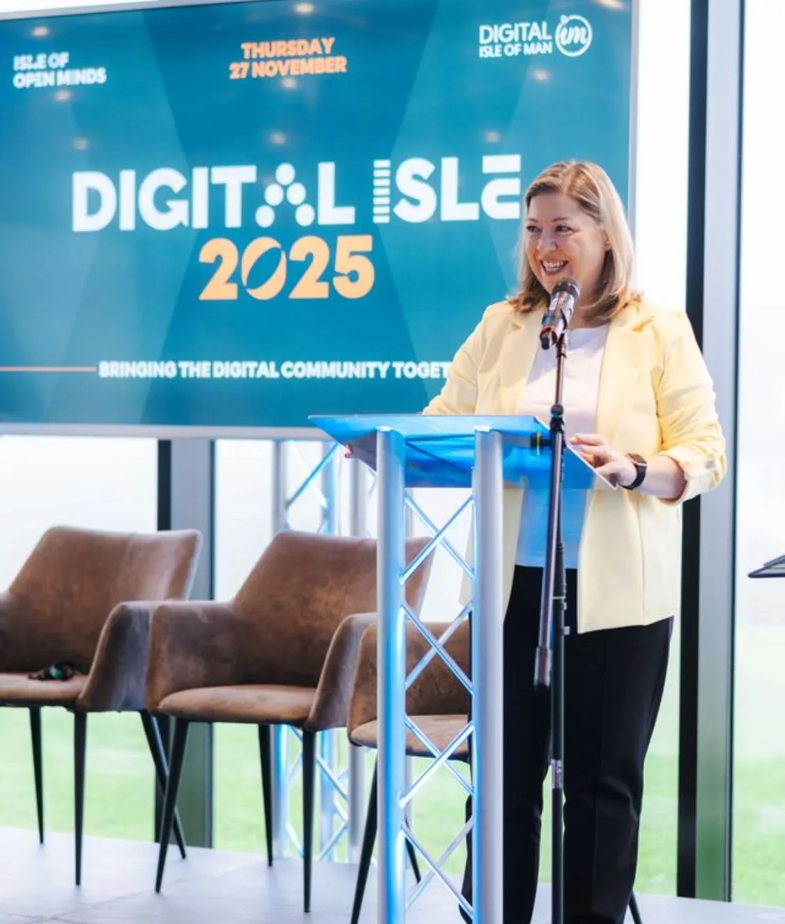 Woman in yellow blazer speaking at a podium with microphone in front of a Digital Isle 2025 event screen.