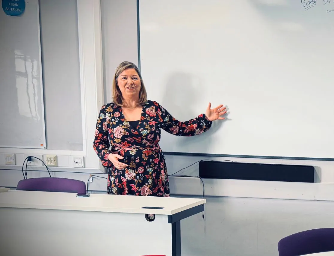 Woman in floral dress standing beside a whiteboard in a classroom, gesturing with her right hand.