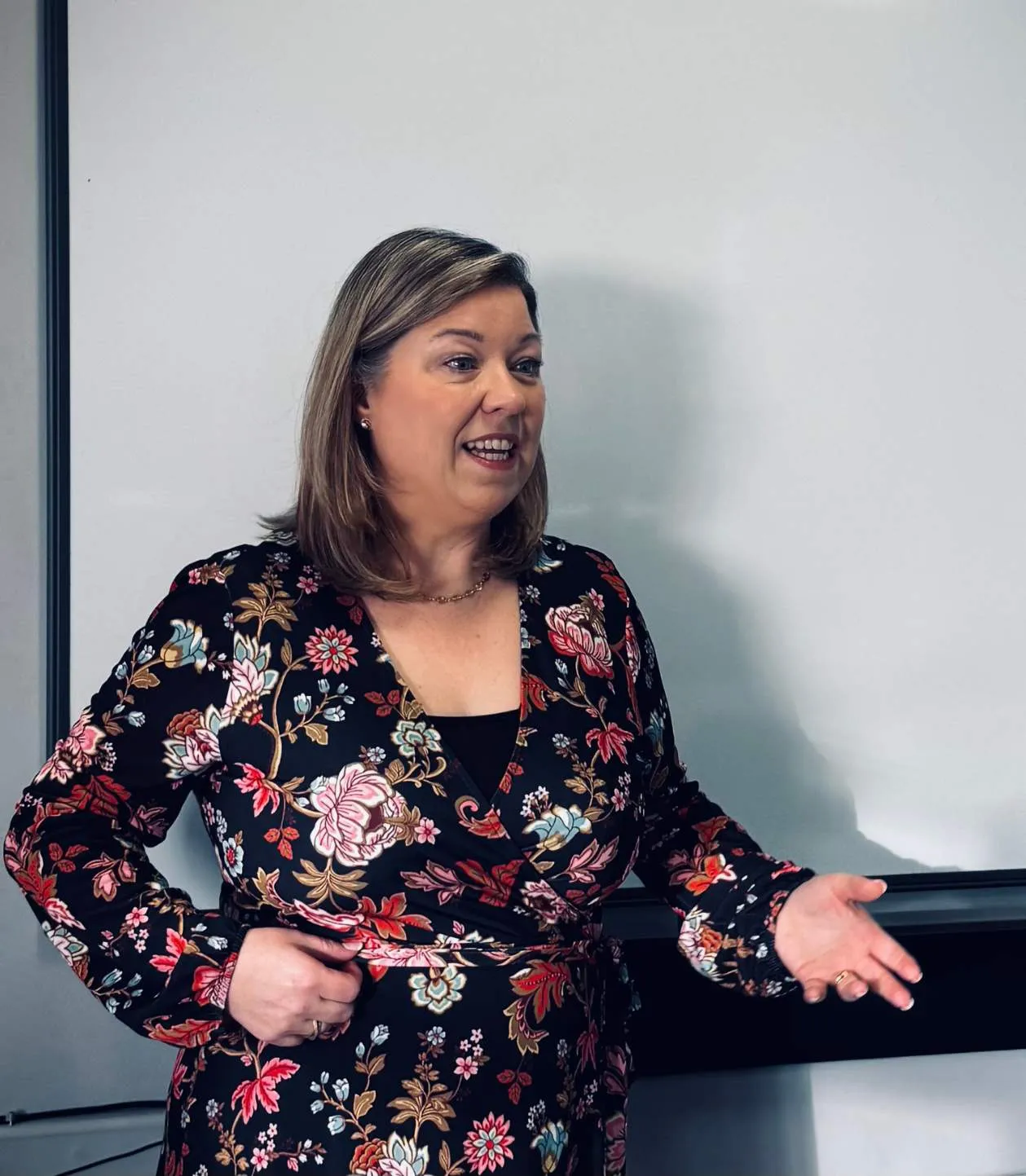 Woman in a floral dress gesturing with her hands while speaking in front of a whiteboard.