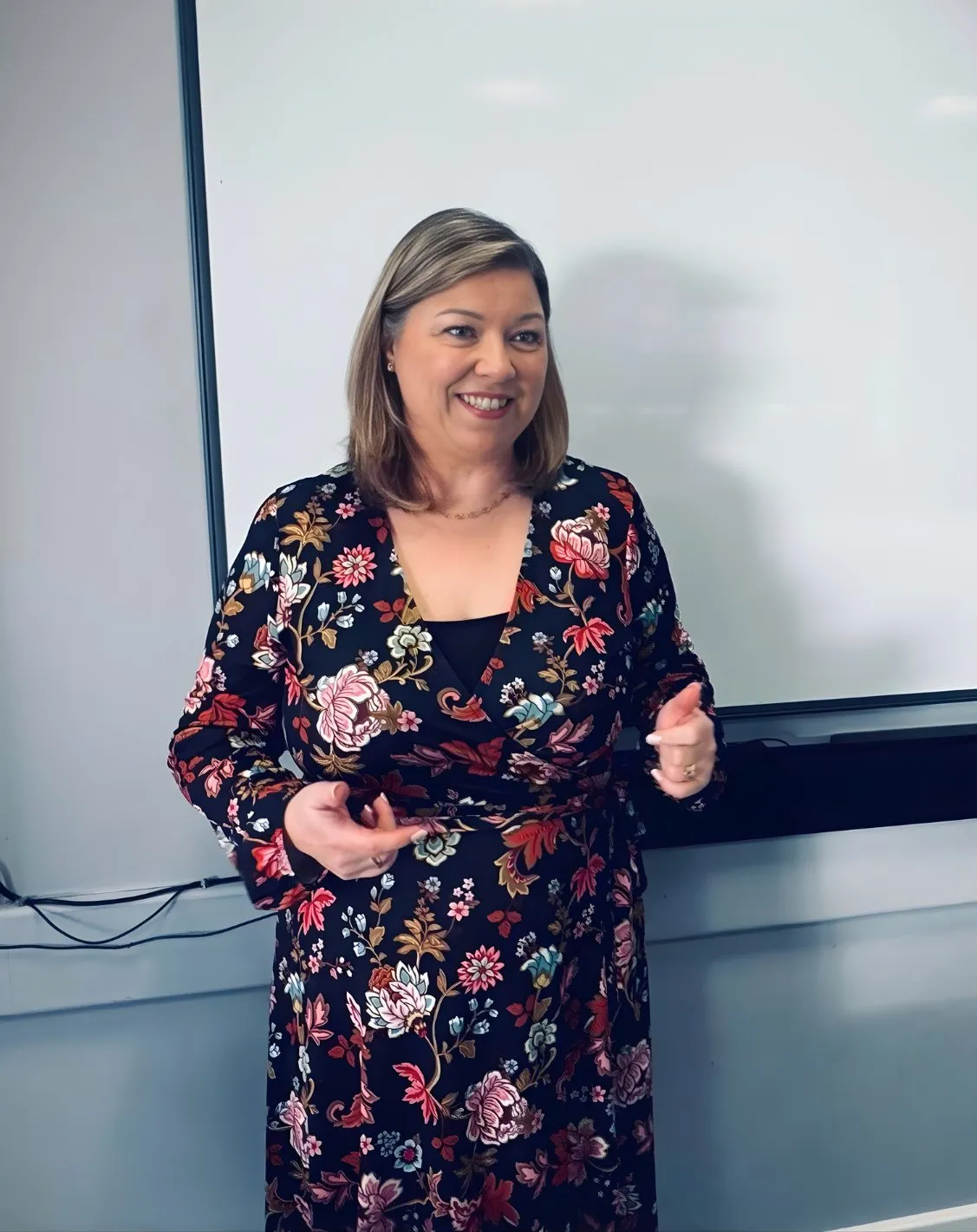 Smiling woman with shoulder-length hair wearing a floral dress standing in front of a whiteboard.