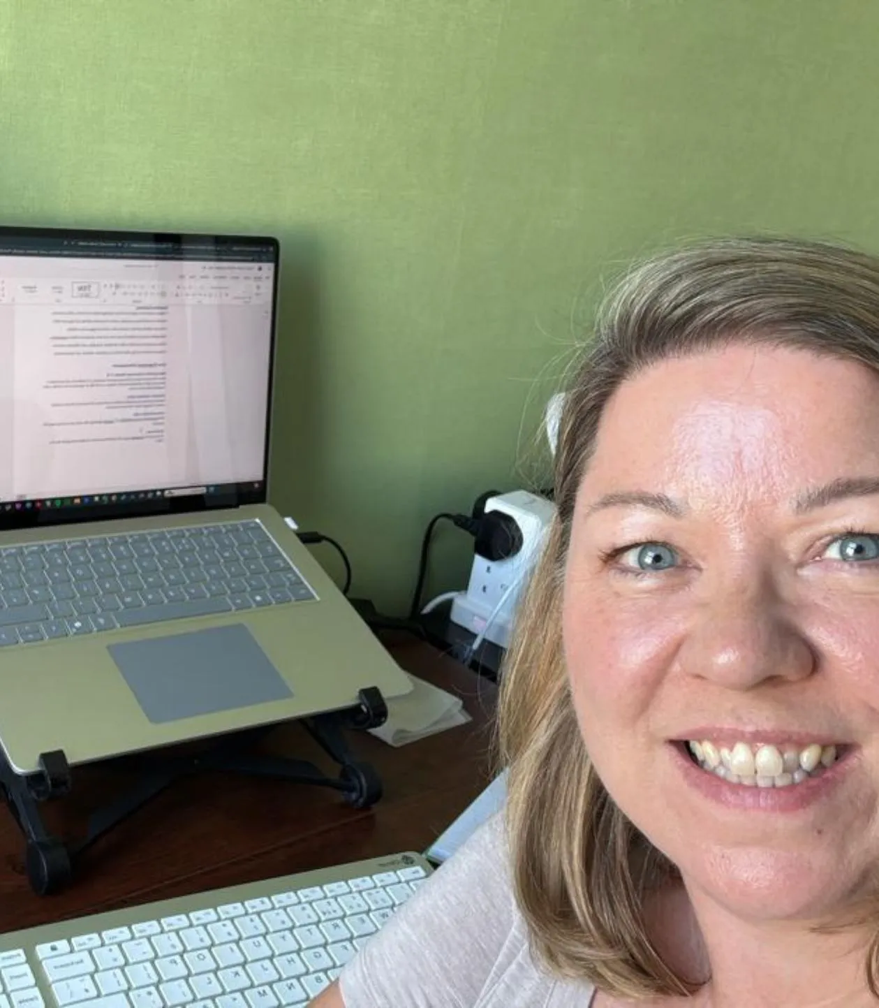 Woman smiling with blue eyes sitting at a desk with a laptop on a stand and a wireless keyboard.