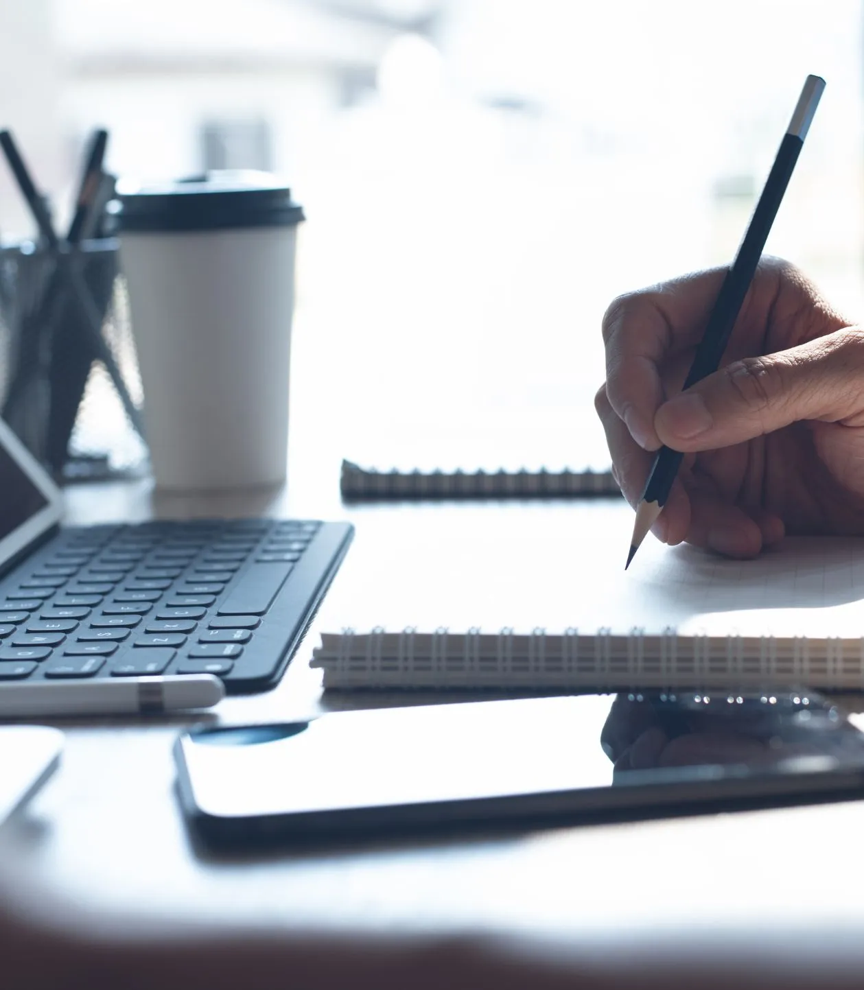 Hand writing with pencil on a spiral notebook beside a keyboard, smartphone, and coffee cup on a desk.