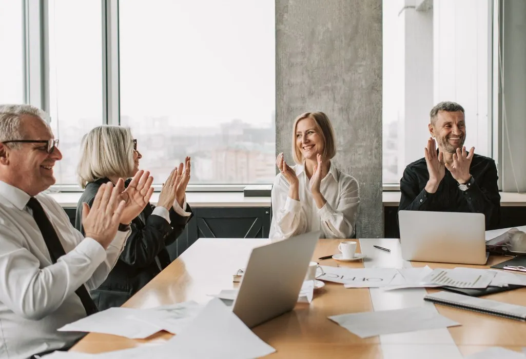 Four diverse business professionals sitting at a conference table clapping and smiling in a modern office.