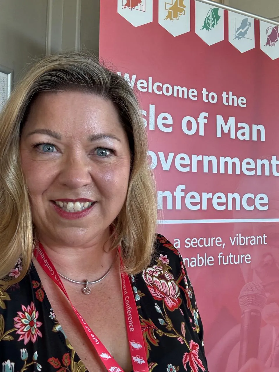 Smiling woman with blonde hair wearing a floral dress and a red lanyard stands in front of a red banner reading 'Welcome to the Isle of Man Government Conference'.