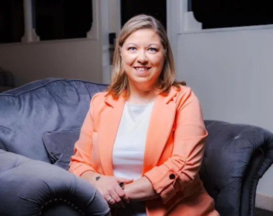 Smiling woman with shoulder-length blonde hair wearing a peach blazer and white shirt sitting on a gray upholstered chair.