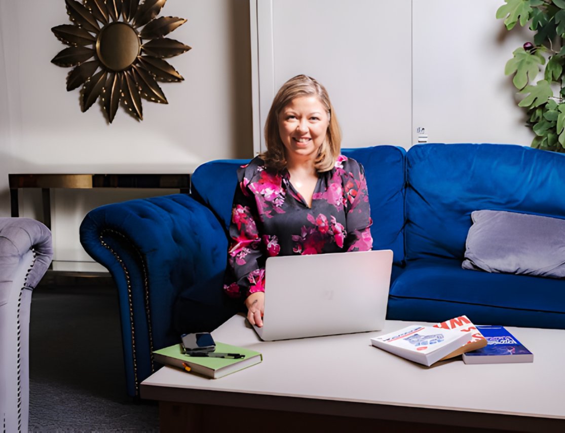 Woman in a black and pink floral blouse sitting on a blue couch working on a laptop with books and a phone on the coffee table.