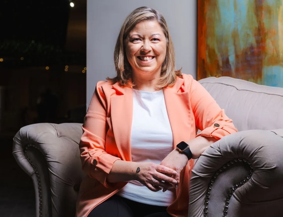 Smiling woman with shoulder-length hair wearing a coral blazer and white shirt sitting on a plush beige armchair.