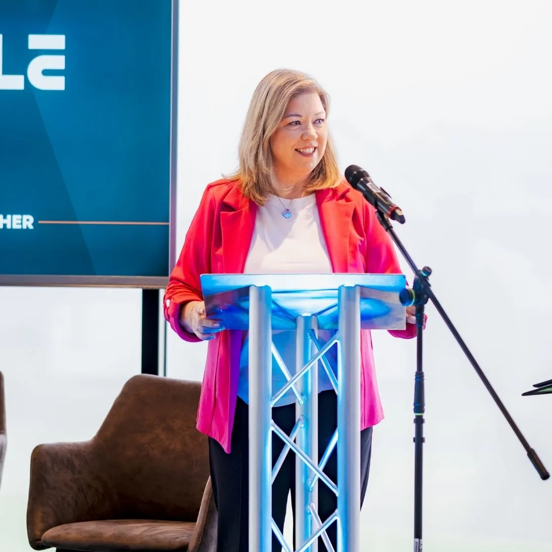 A woman in a red blazer stands smiling behind a podium with a microphone, with a brown chair and a screen partially visible in the background.
