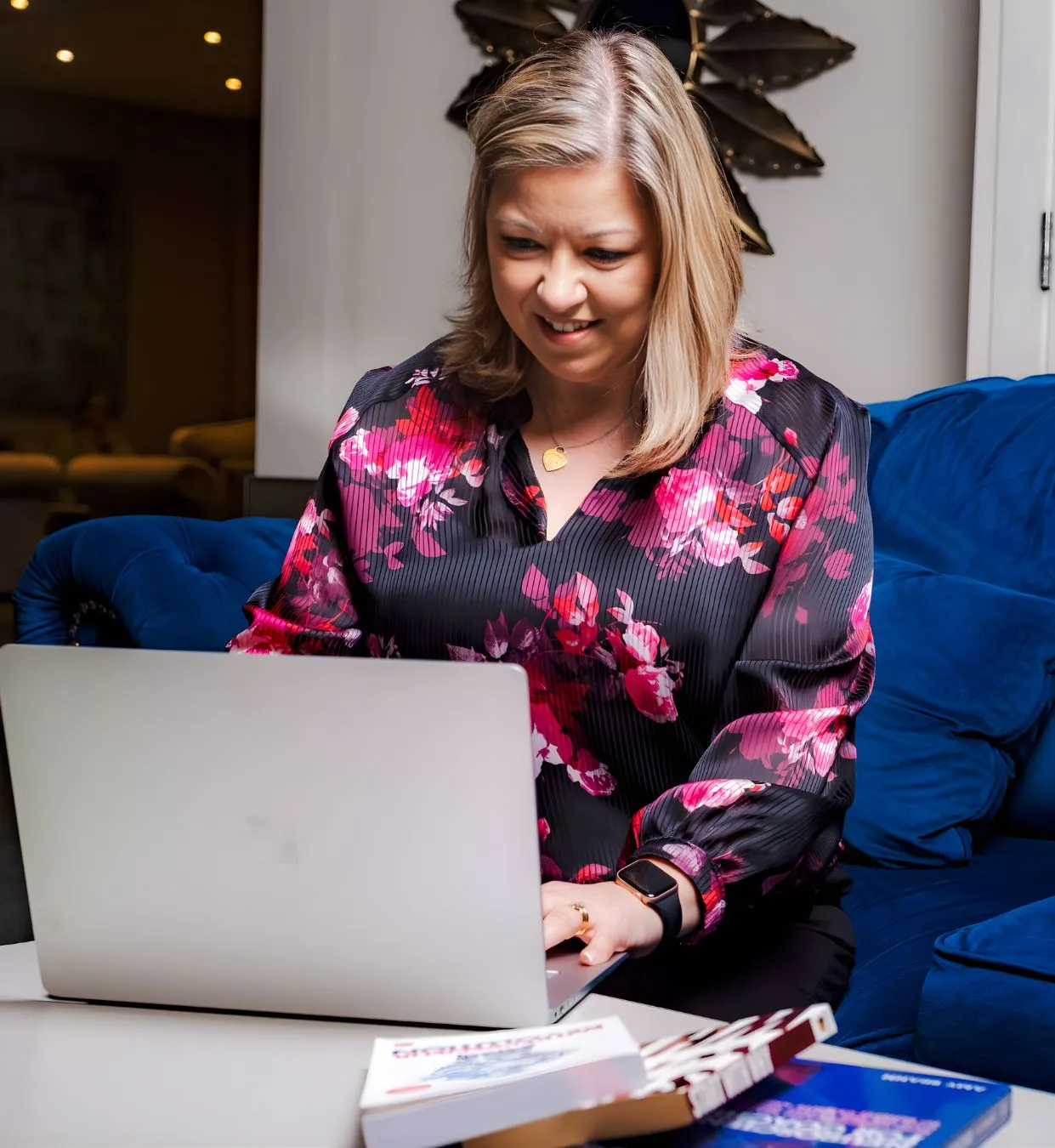 Woman with blonde hair sitting on a blue couch, working on a laptop at a white table with books.