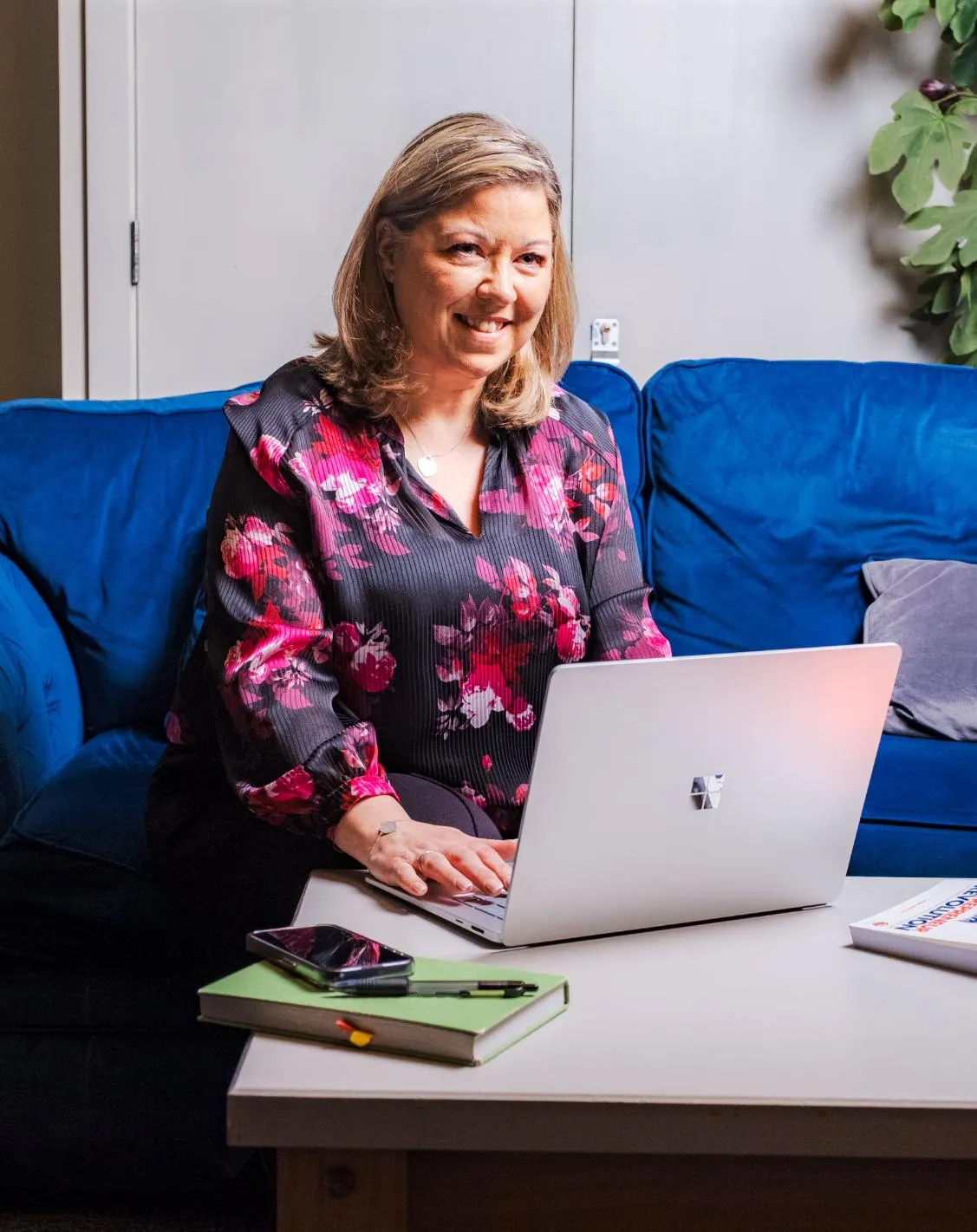 Smiling woman in a floral blouse sitting on a blue couch working on a silver laptop at a white table with a phone and notebook.