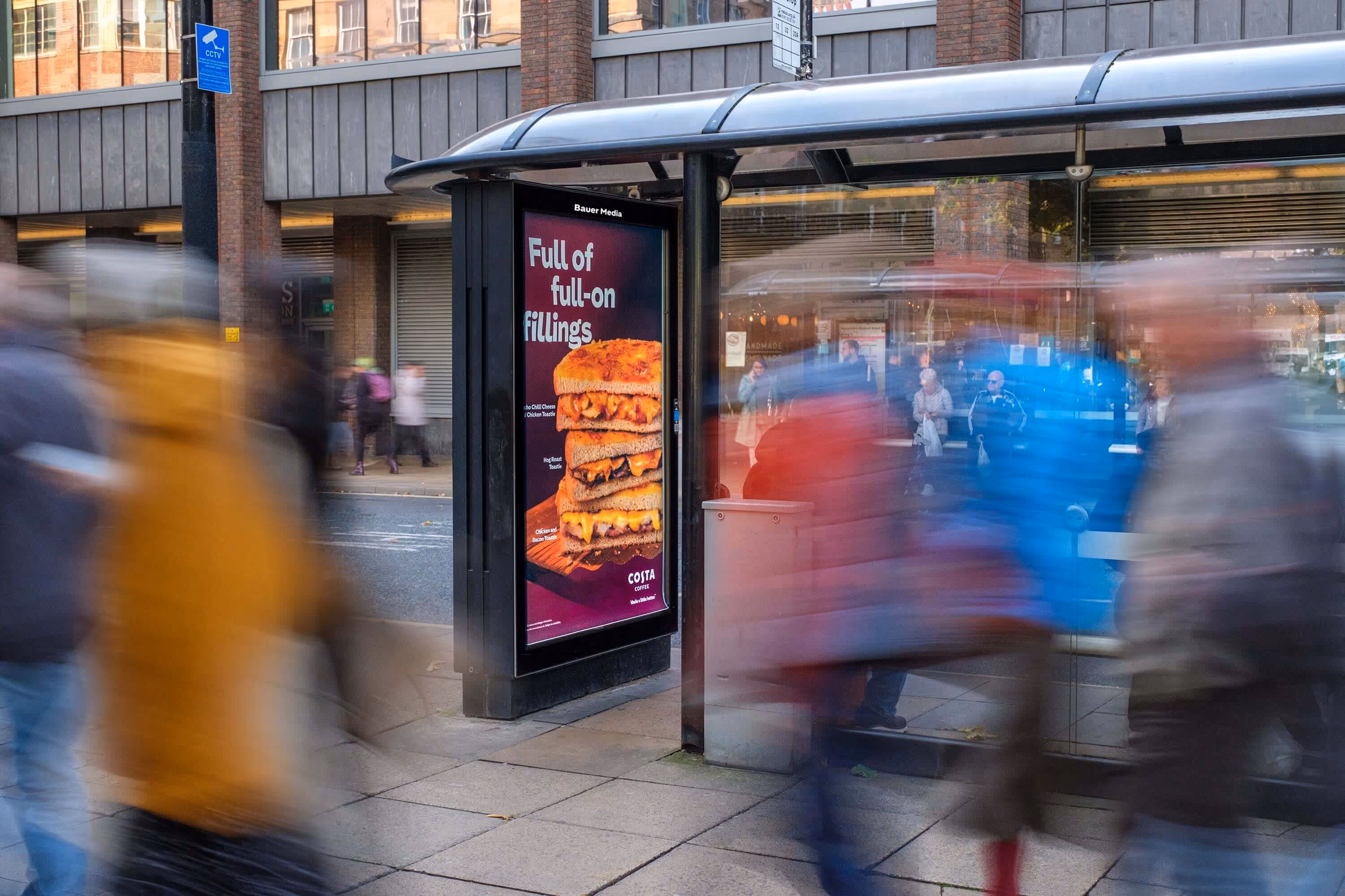 Bauer Media Outdoor digital advertising screen at a busy bus stop displays a Costa ad featuring stacked toasties with the tagline “Full of full-on fillings,” as pedestrians pass by.