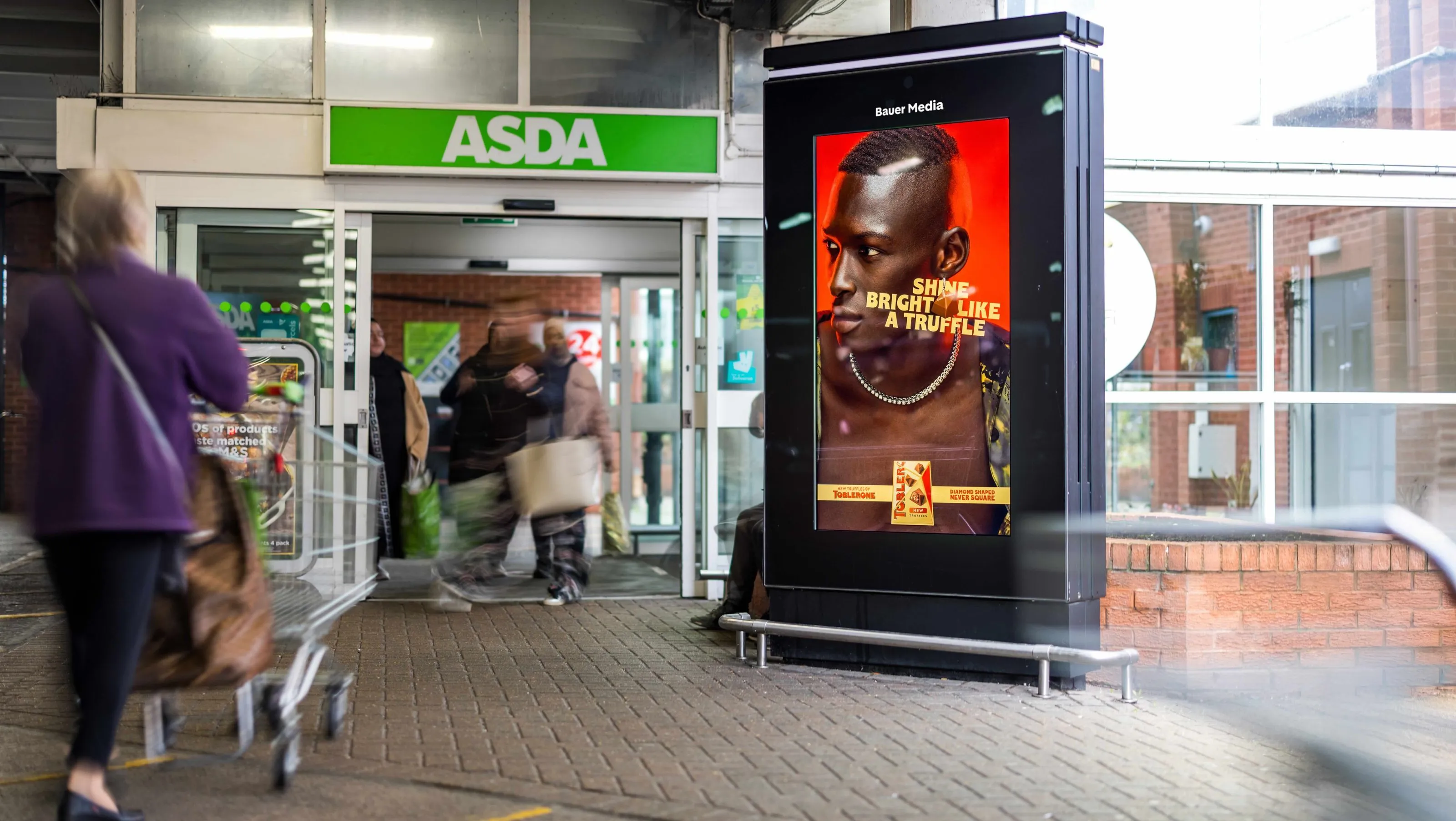 Bauer Media Outdoor digital advertising screen outside an ASDA supermarket displays a Toblerone ad featuring a model with the headline “Shine Bright Like a Truffle". 