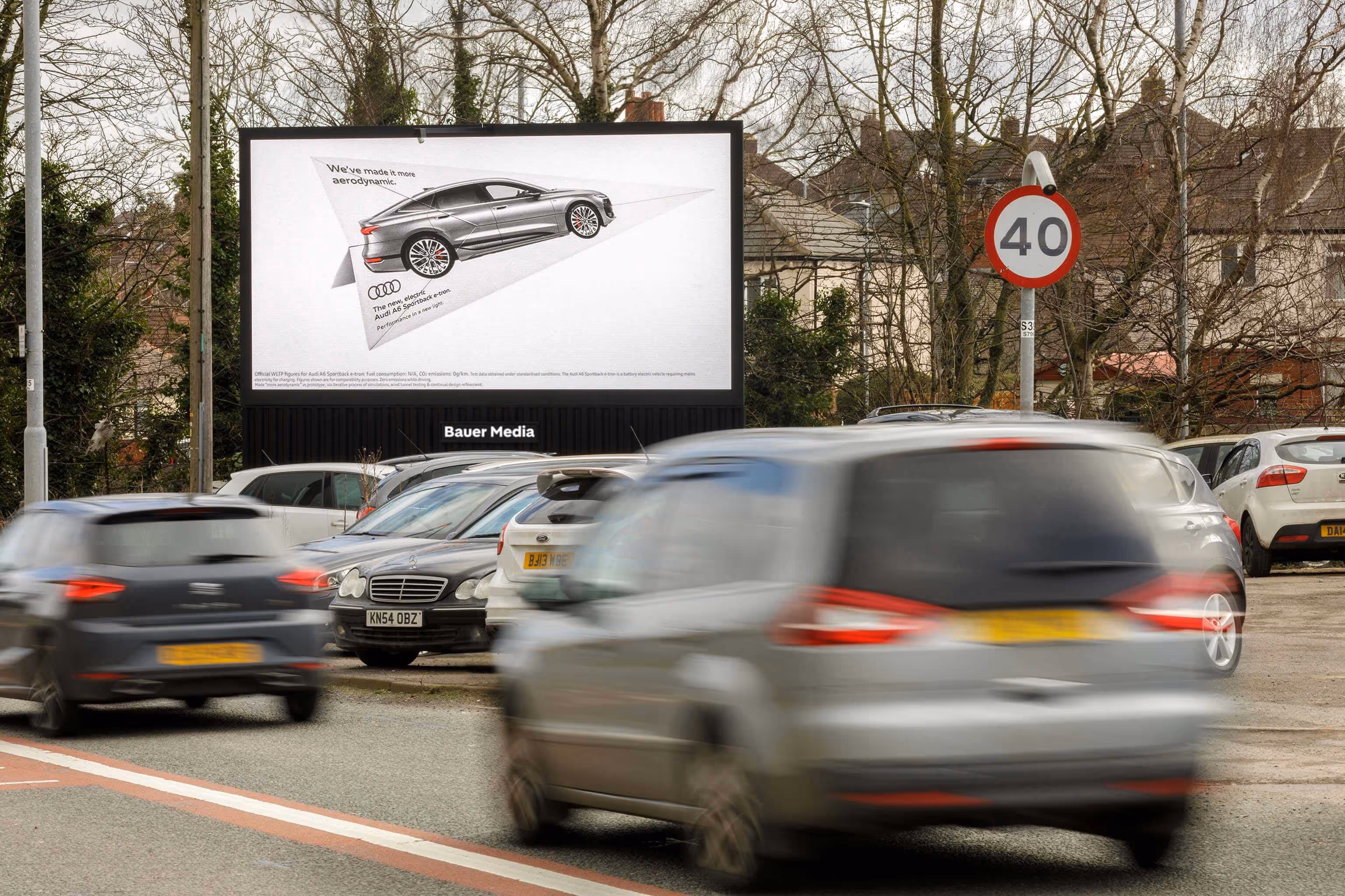 Bauer Media Outdoor roadside digital billboard displays a sleek Audi ad in a residential area, as cars drive past and queue in traffic beneath a 40 mph speed limit sign.
