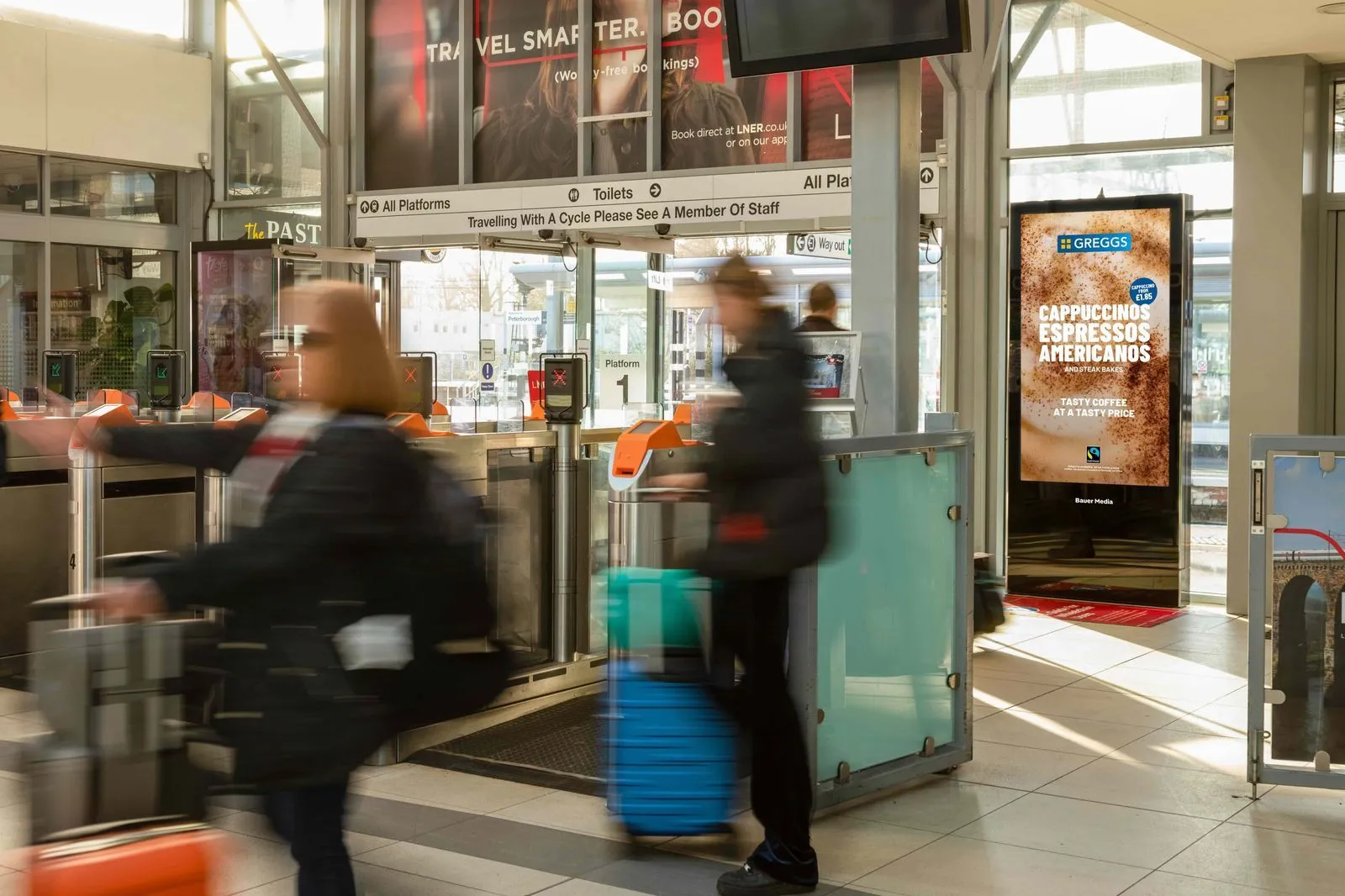 Bauer Media Outdoor digital advertising screen near ticket barriers in a train station displays a Greggs coffee ad promoting cappuccinos, espressos, and americanos, as travelers move through with luggage.