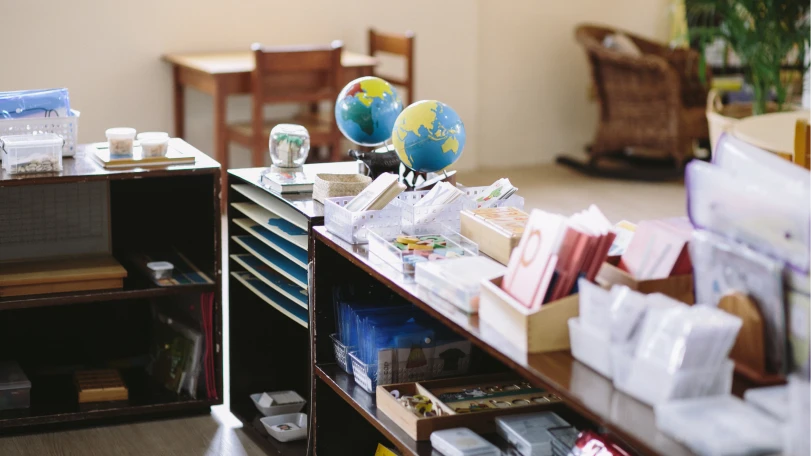 Classroom shelves with educational materials, including globes, books, and various learning tools, with a wooden table and chairs in the background.
