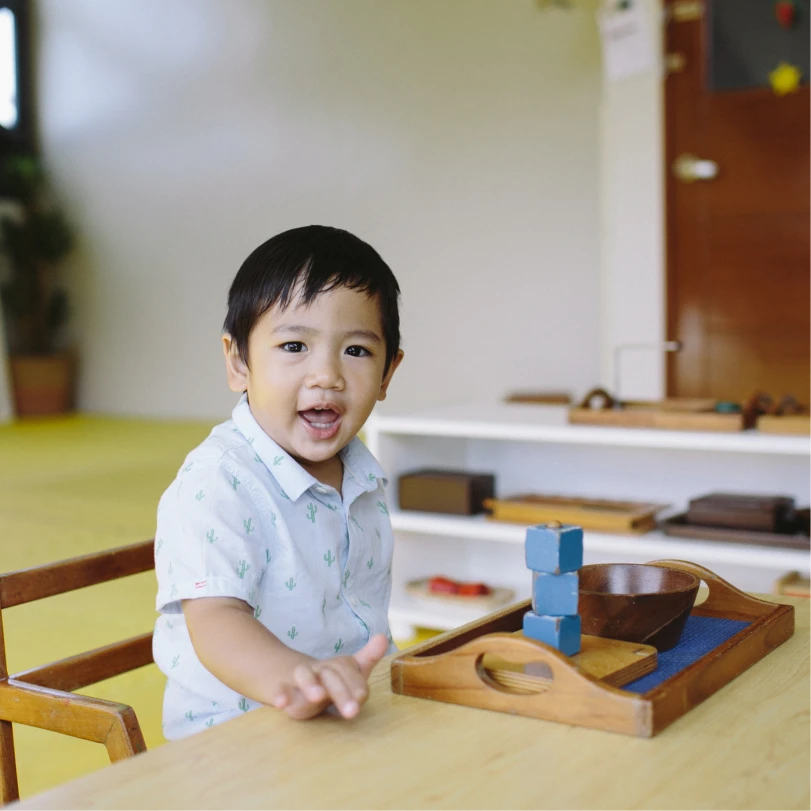Young boy sitting at a table smiling with a wooden tray holding blue blocks and a brown bowl in front of him.