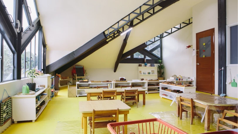 Bright classroom with slanted ceiling, wooden tables and chairs, low shelves with educational materials, and large windows on the left.