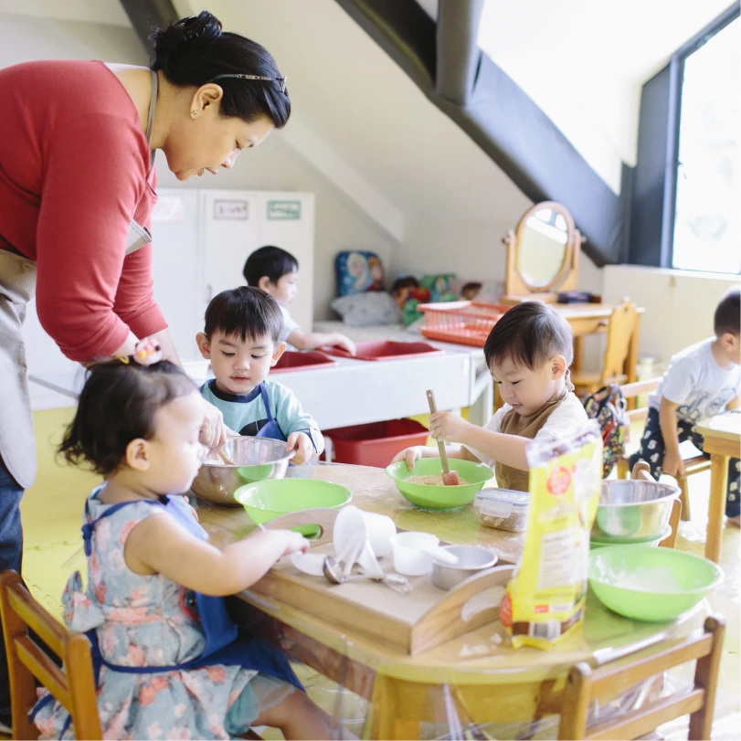 Teacher assisting three young children at a table as they mix ingredients in a bright classroom.