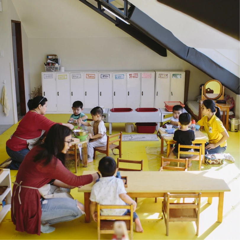 Two women interacting with young children seated at small tables in a bright classroom with yellow flooring and white storage cabinets.