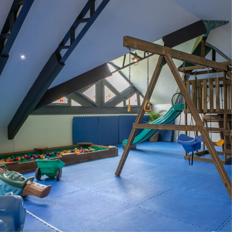 Indoor children's playroom with blue padded floor, wooden swing set, slide, ball pit, and toys under a slanted roof with windows.