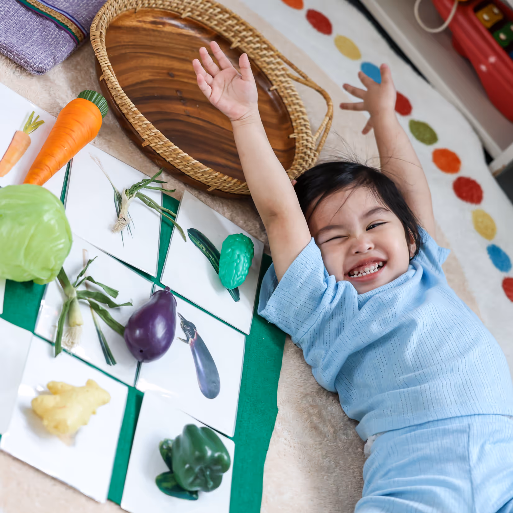 Smiling child lying on the floor with arms raised next to toy vegetables and cards with vegetable images.