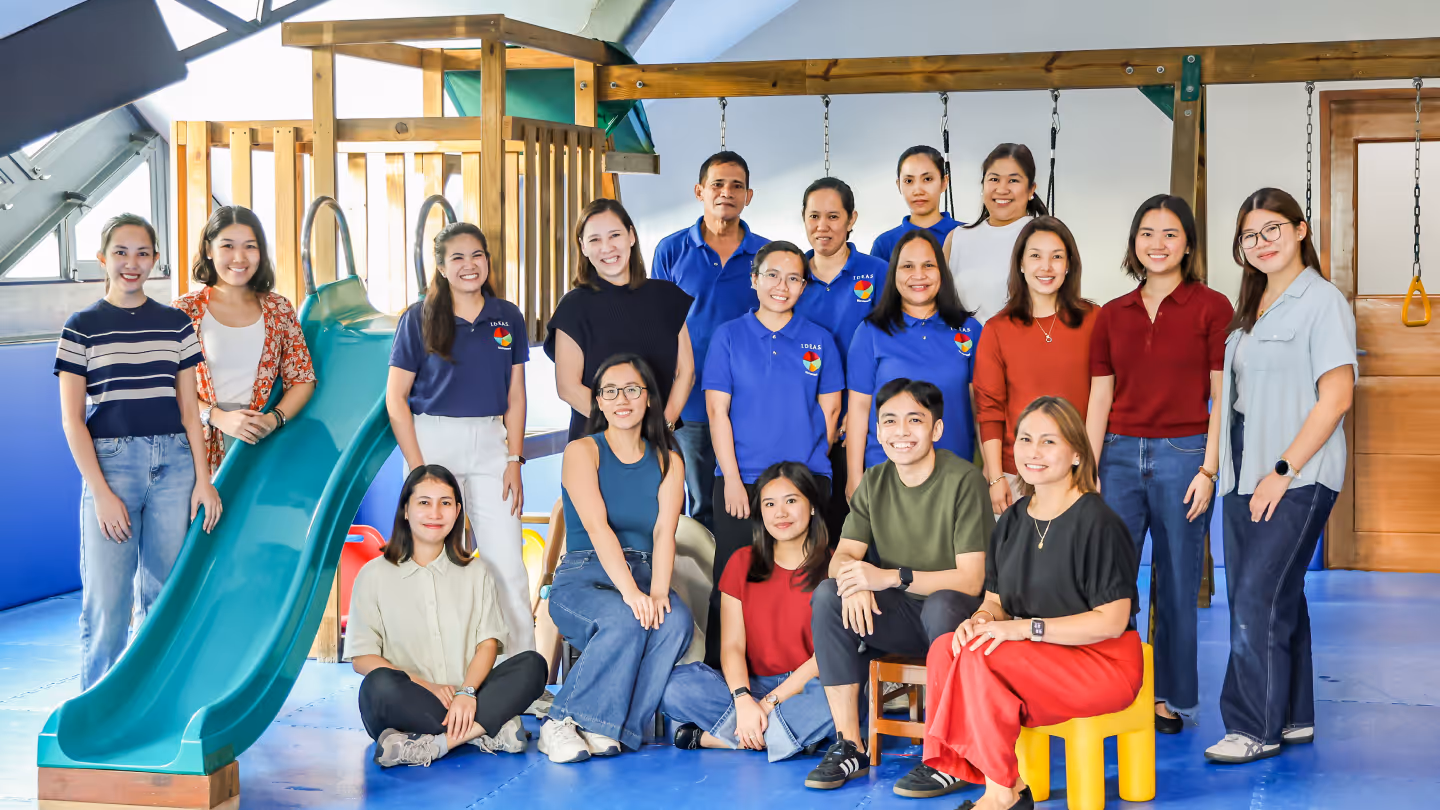 Group of 17 smiling adults posed together indoors near a green slide and wooden play structure.