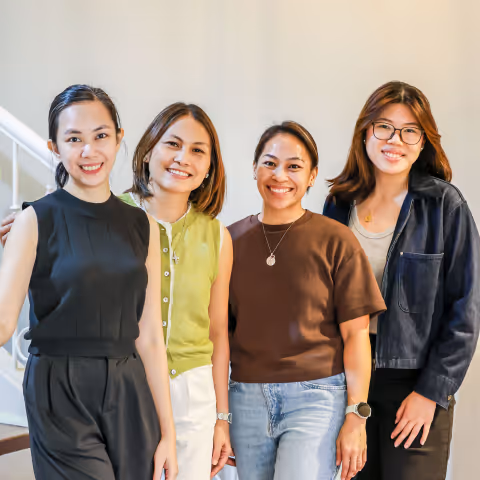 Four smiling women standing indoors against a light wall, dressed casually and looking at the camera.