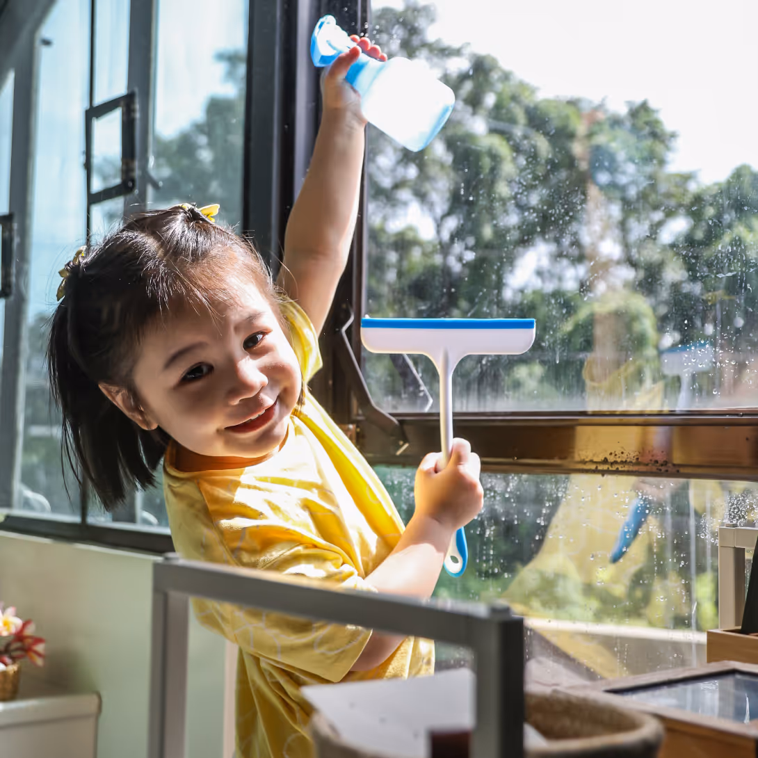 A young girl smiling while cleaning a window with a spray bottle and squeegee.