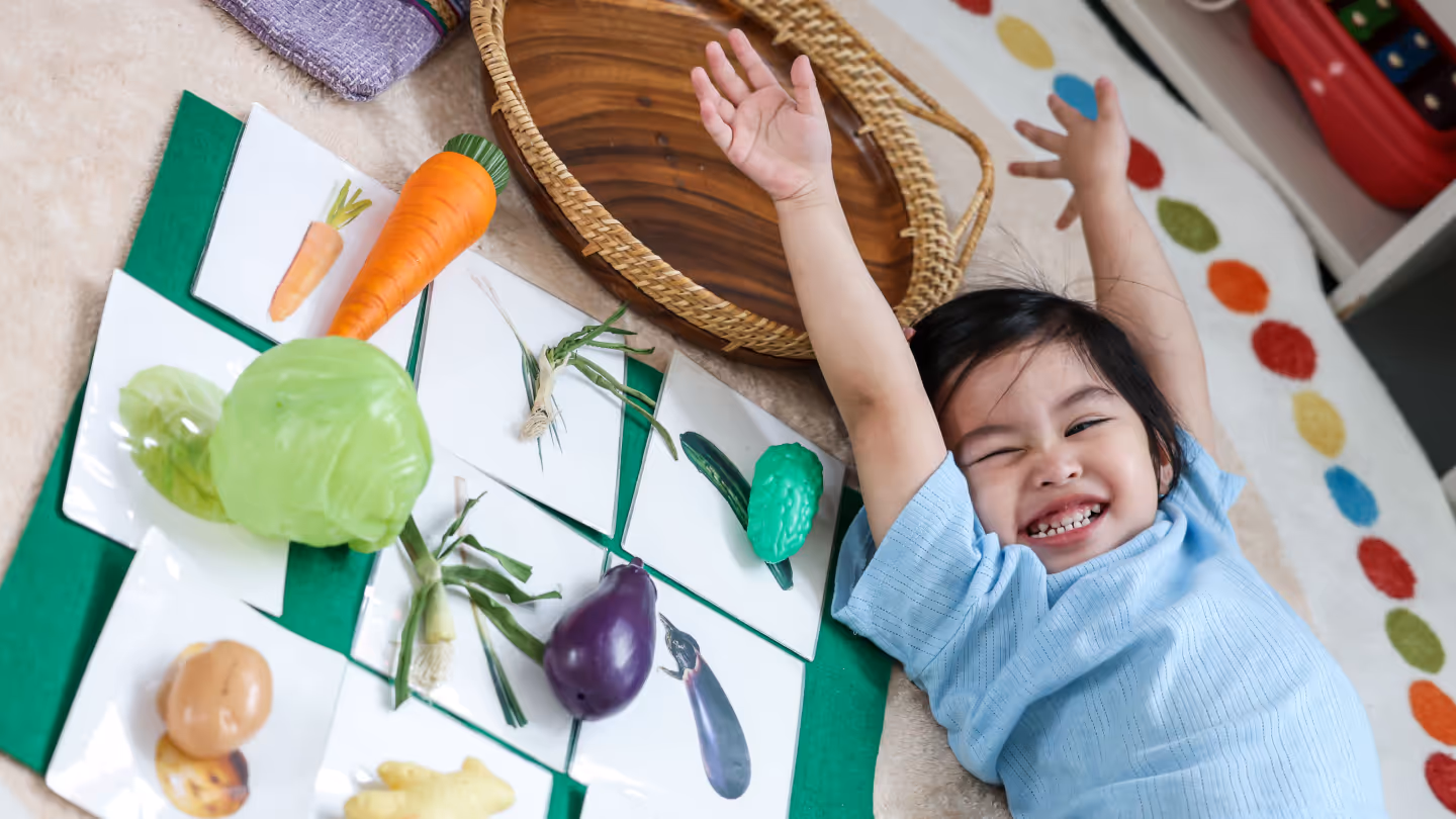 Smiling child lying on the floor next to flashcards and toy vegetables arranged on a green mat.