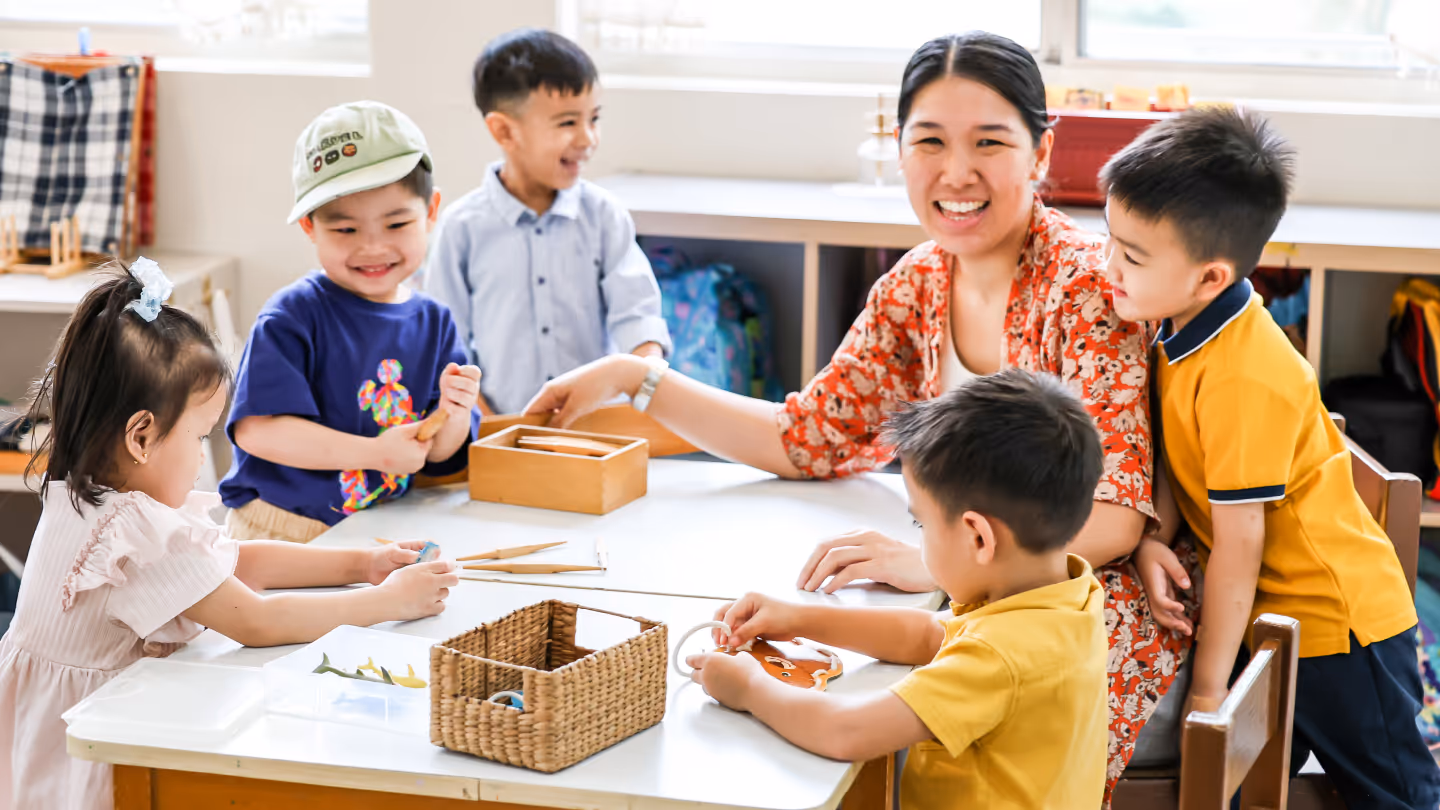 Teacher smiling and interacting with five young children around a table with educational toys in a classroom.