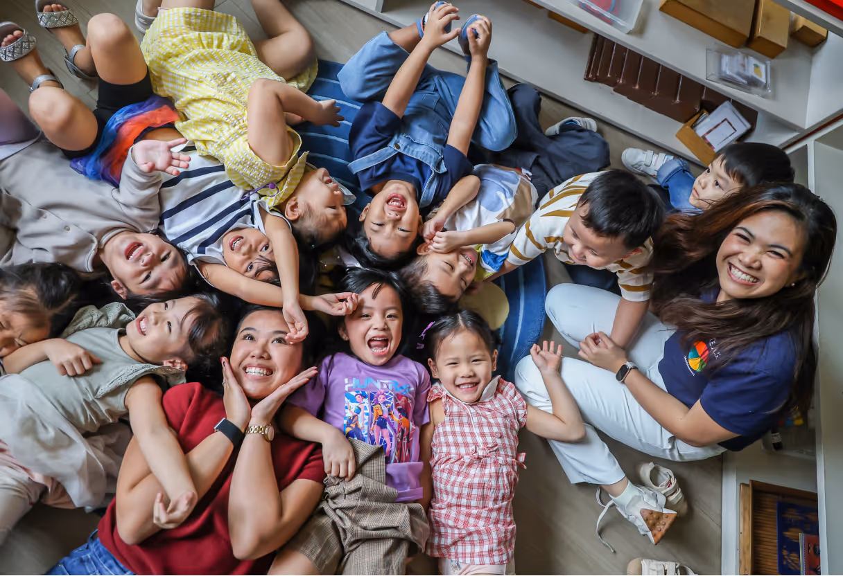 Group of smiling children and two women lying on the floor in a circle, enjoying a playful moment indoors.