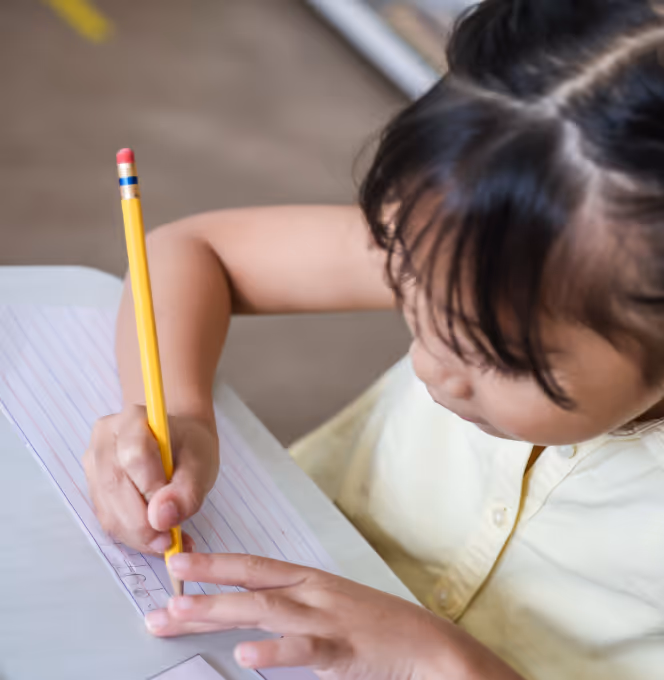 Child writing with a yellow pencil on lined paper at a desk.
