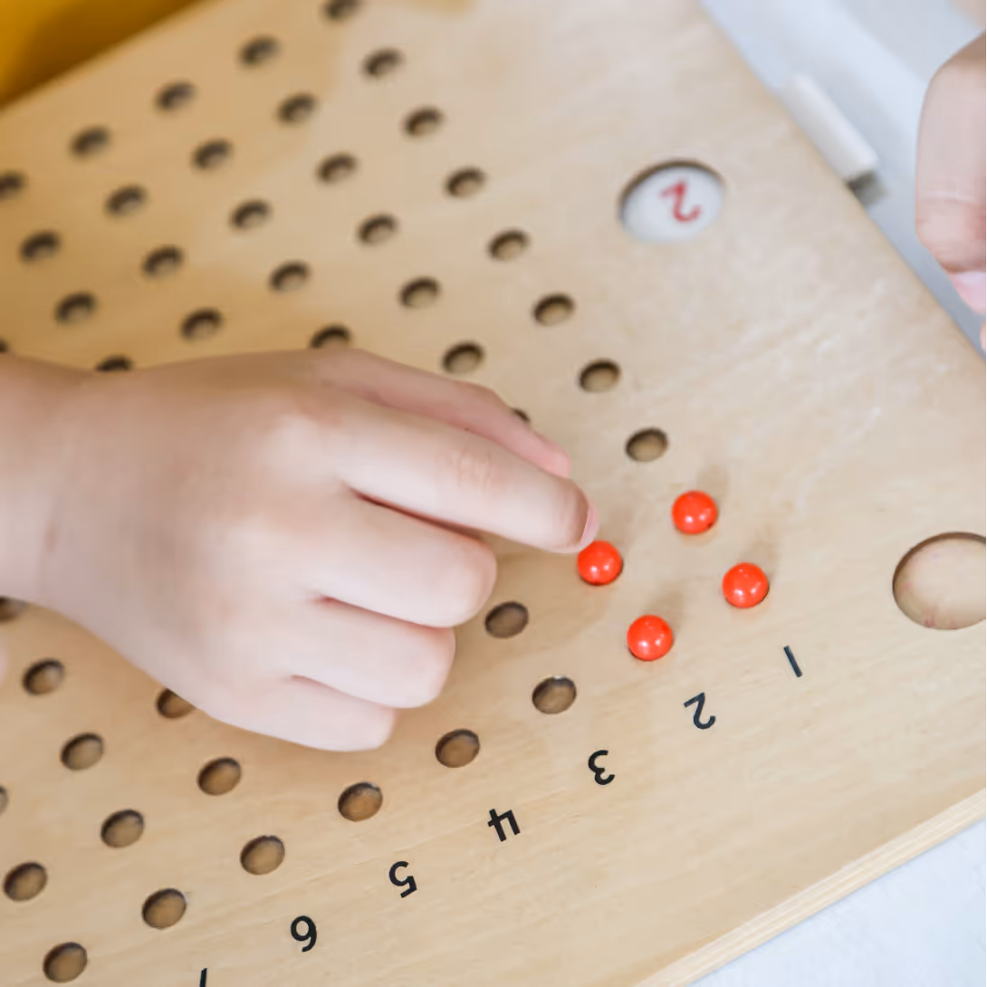 Child's hand placing red beads on a wooden Montessori number board near numbers 1 to 6.