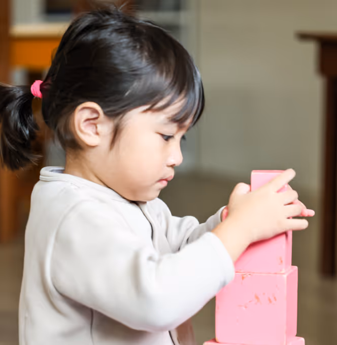 Young girl with a ponytail focused on stacking pink wooden blocks indoors.