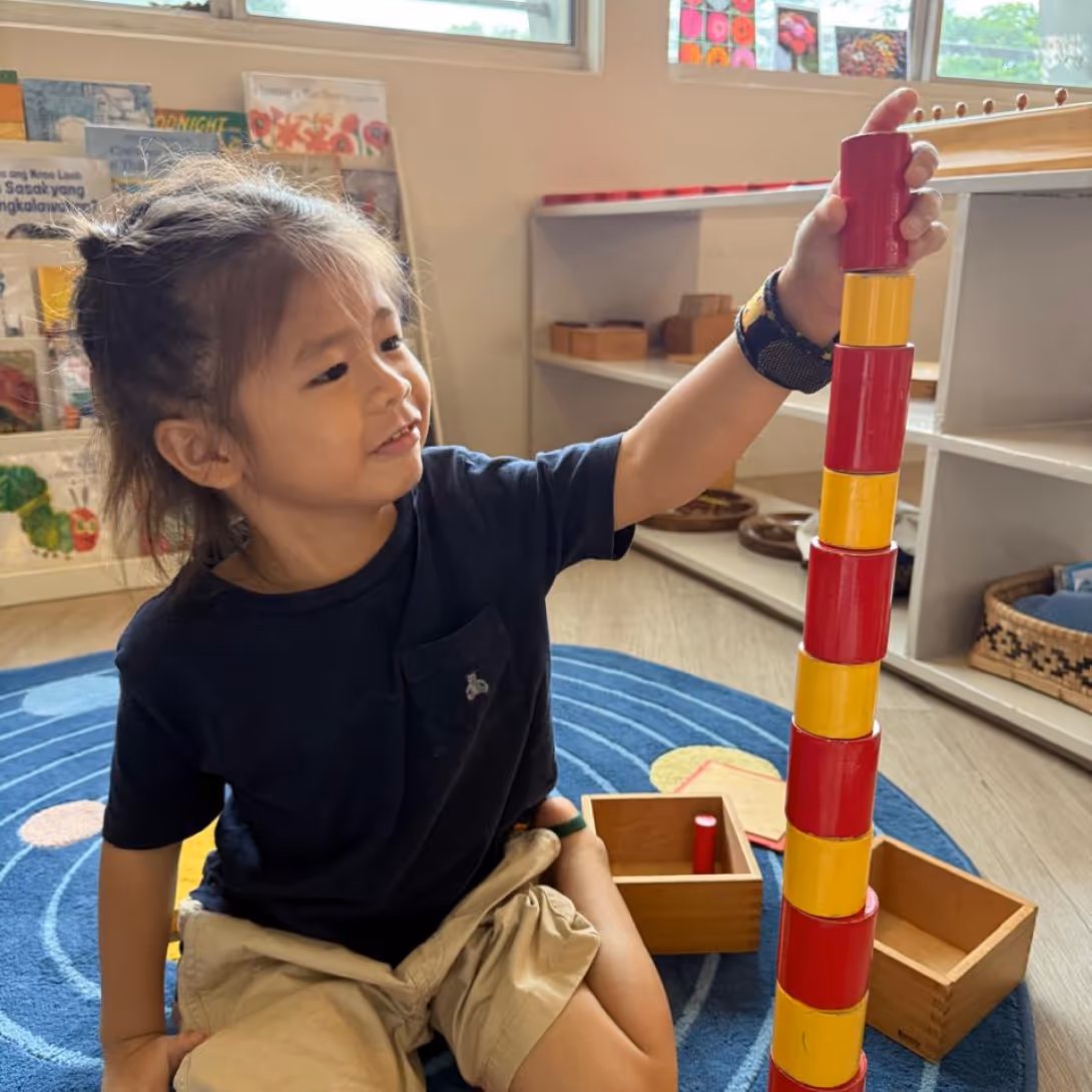 A child sitting on a blue circular rug stacking red and yellow cylindrical blocks in a classroom setting.