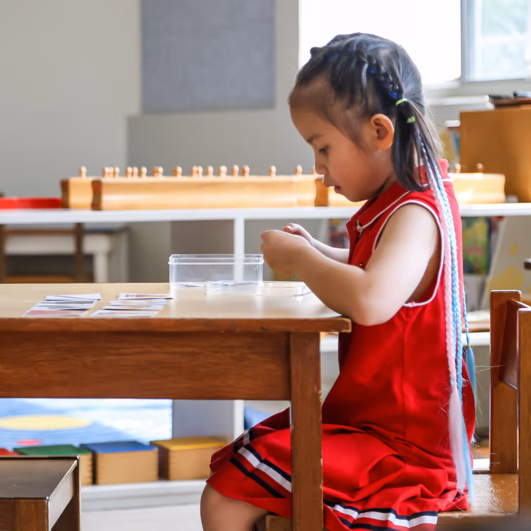 Young girl in a red dress sitting at a wooden table, focused on small cards in front of her in a classroom setting.