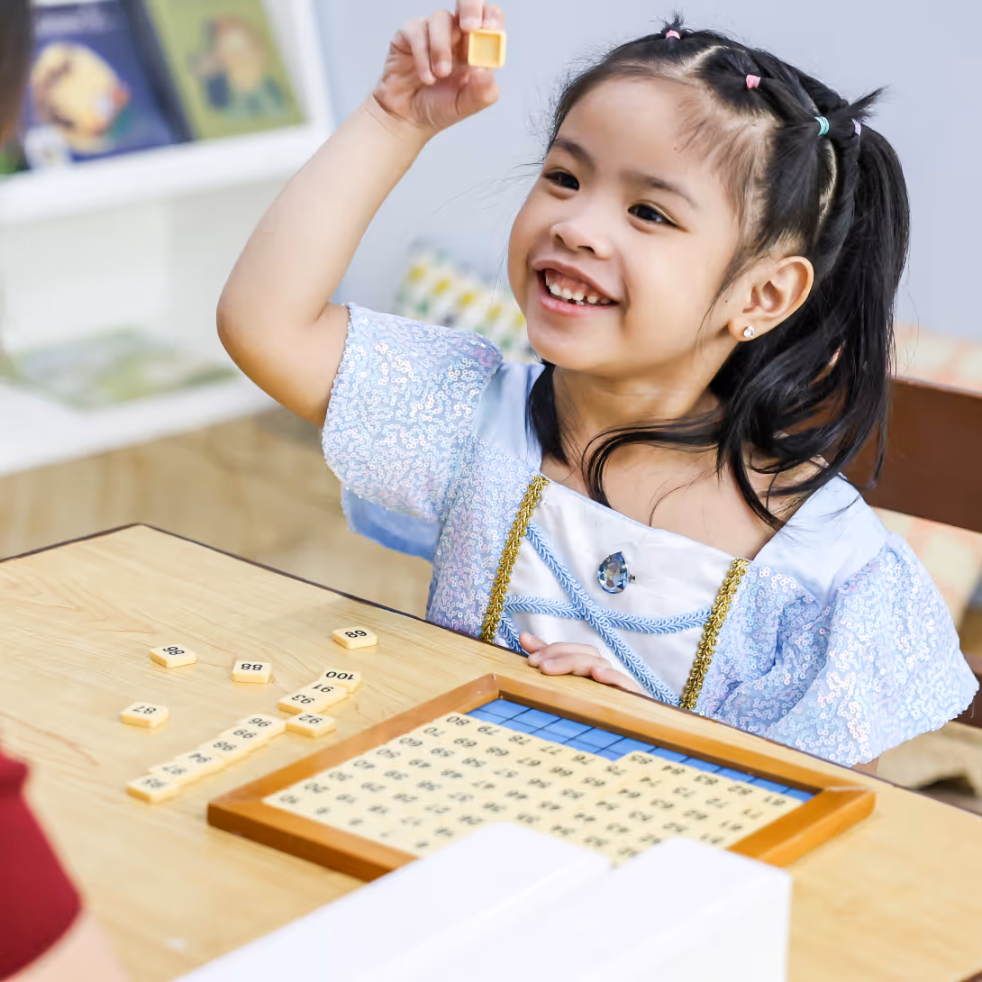 Smiling young girl in a blue dress holding a number tile above a wooden math board game with scattered tiles.