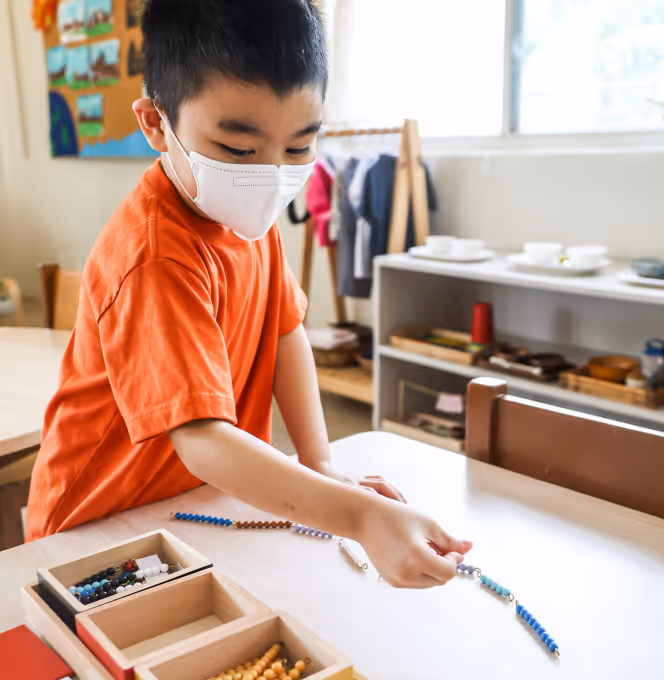 Child wearing a white face mask and orange shirt arranging colorful bead chains on a table in a classroom.