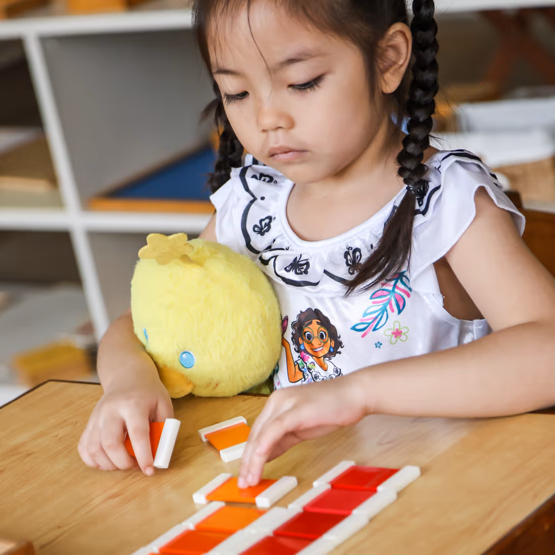 Young girl with braided hair holding a yellow plush chick while playing with colorful tiles on a wooden table.