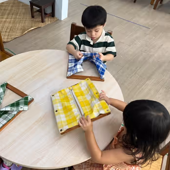 Two children sitting at a round table playing with fabric folding boards featuring blue and yellow plaid cloths.
