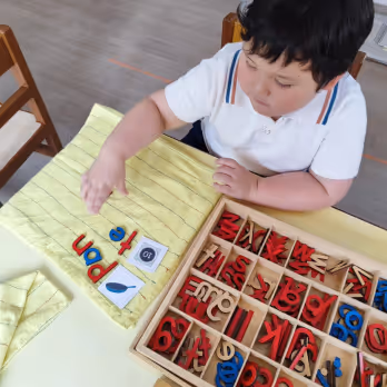 Child arranging colorful wooden letters to form words on a yellow cloth next to a box of more letters.