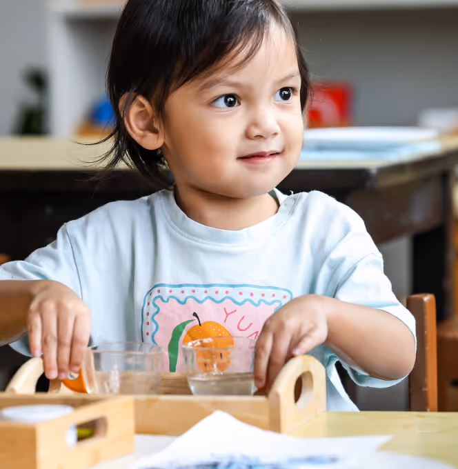 Young child sitting at a table holding a wooden tray with glass cups, looking to the side and smiling slightly.