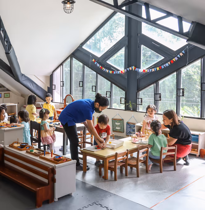 Teacher assisting young children engaged in various learning activities at tables inside a bright classroom with large windows and colorful decorations.