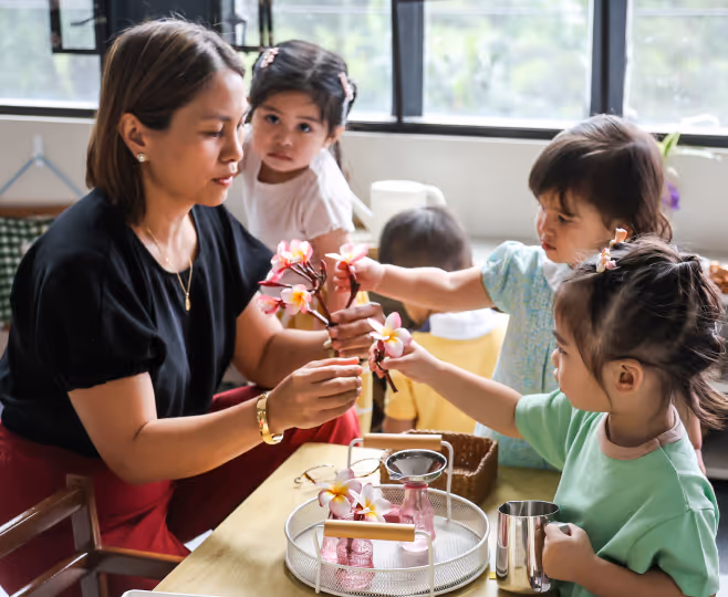 Woman and three young children seated at a table, arranging pink and white flowers in small vases.