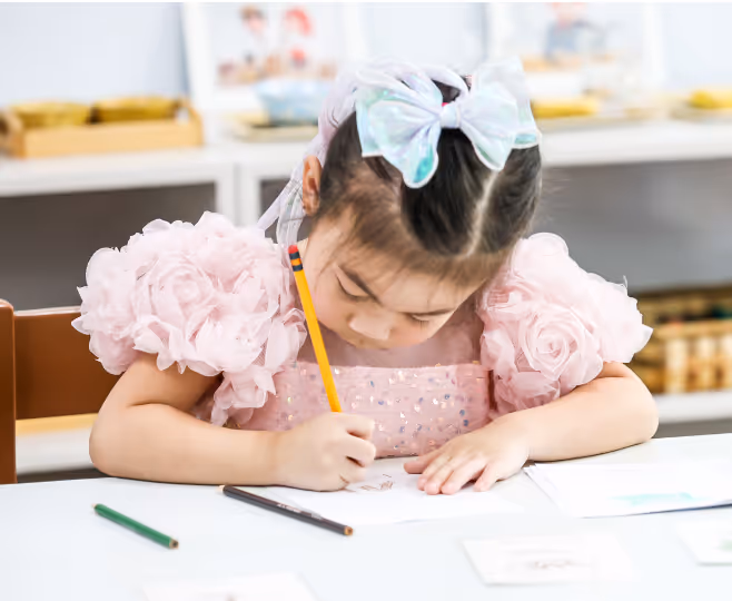 Young girl with a white bow and pink puffy dress concentrating on drawing with a pencil at a white table.