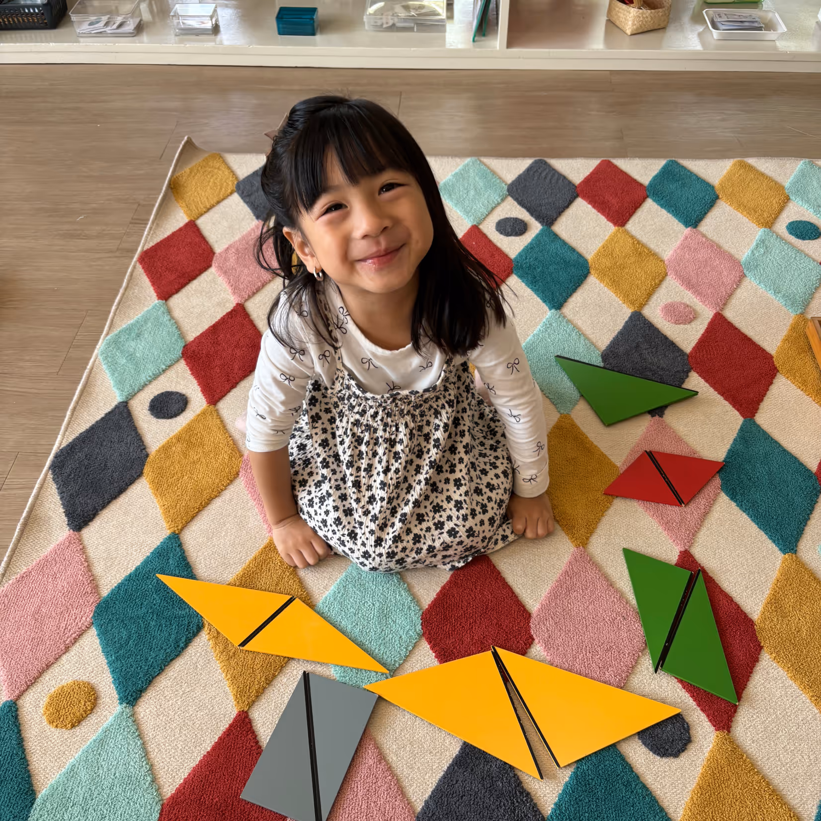 Smiling young girl sitting cross-legged on a colorful diamond-patterned rug with large yellow, green, red, and gray geometric shapes scattered around her.