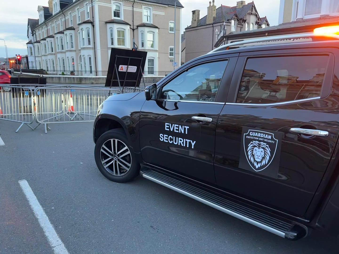 Black security vehicle with 'Event Security' and lion logo parked near metal barricades on a street in front of residential buildings.